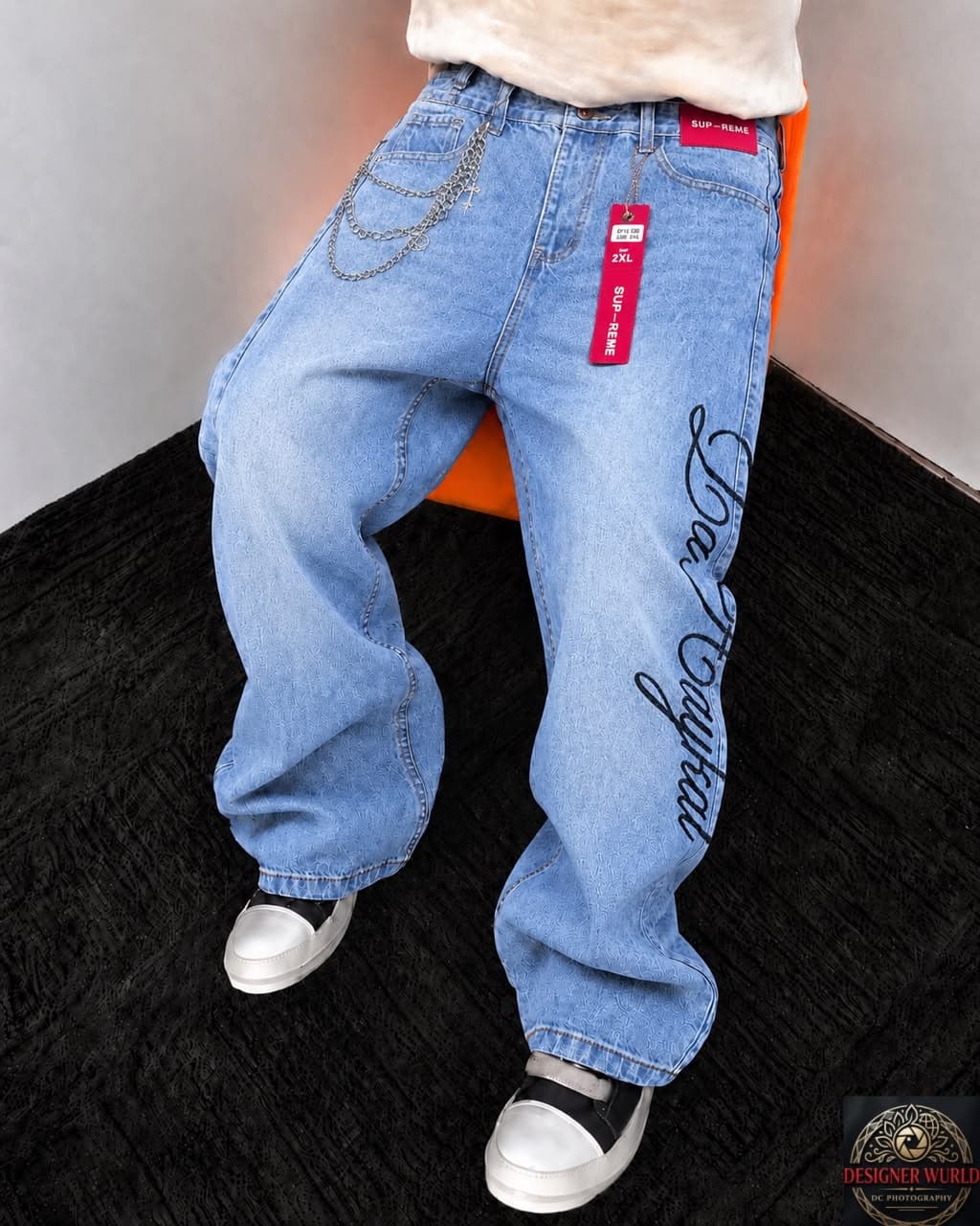 Two tan baggy jeans laid on asphalt with skateboard: one with back embroidery and patches, one with front rips and studs.