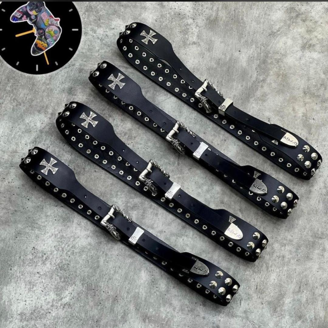 Four black leather belts with studs, cross emblems and varied buckles arranged on grey surface.