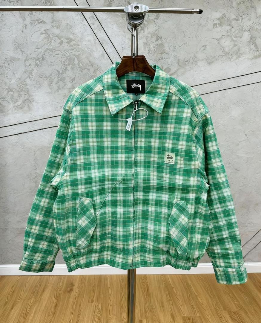 Green checkered Stussy jacket hanging on display stand, front view showing plaid pattern and pockets.