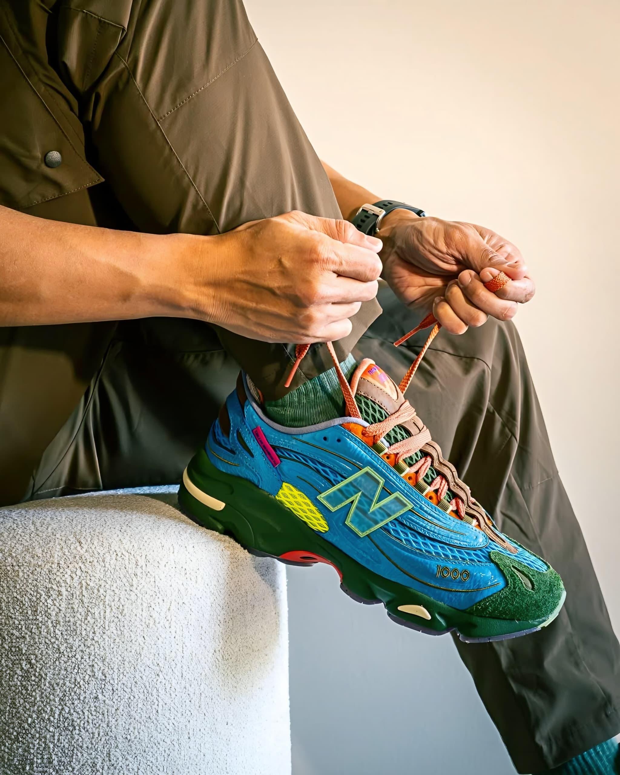 Blue green and orange New Balance 1000 sneakers on foot with hands tying laces.